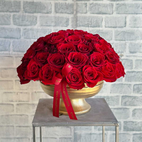 Round arrangement of red roses in a gold pedestal bowl with a red ribbon