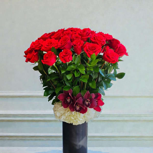 Red roses arranged in a tall black vase with white and burgundy flowers at the base