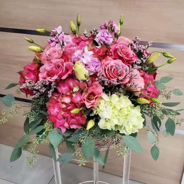 Large pink and cream flower arrangement with roses and hydrangeas in a low basket on a clear stand