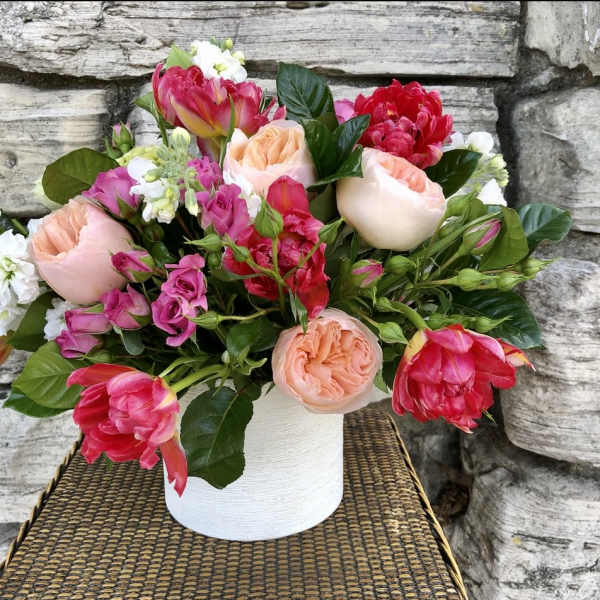 Low arrangement of pink and peach roses, tulips, and white stock in a white ceramic vase