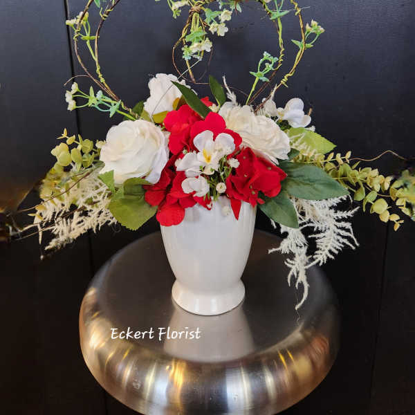 Red and white floral arrangement in a white vase with heart-shaped twig accents
