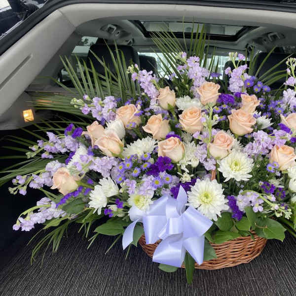 Basket arrangement of peach roses, white daisies, and purple flowers with a lavender bow