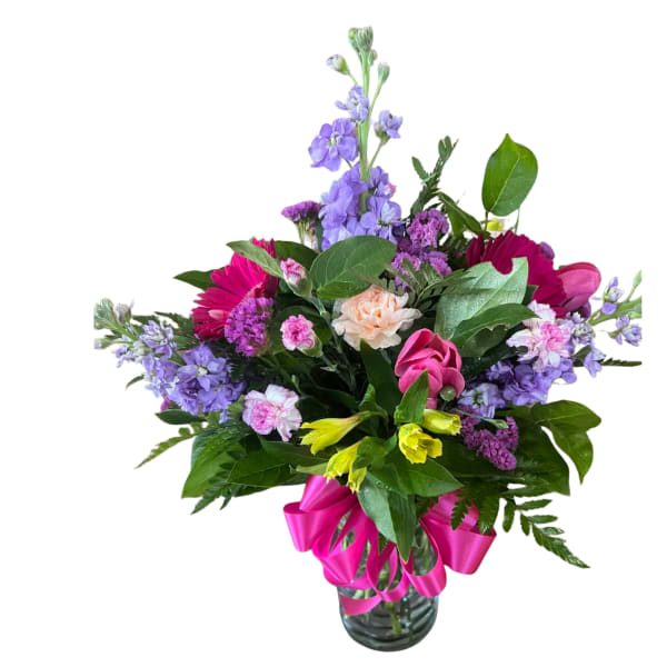 Mixed bouquet of pink, purple, yellow, and peach flowers in a clear glass vase with a bright pink ribbon bow