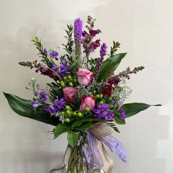 Tall arrangement of pink roses and purple flowers in a clear glass vase with decorative bow