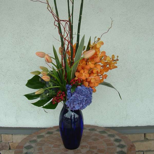 Tall floral arrangement in a blue vase with orange orchids and blue hydrangea