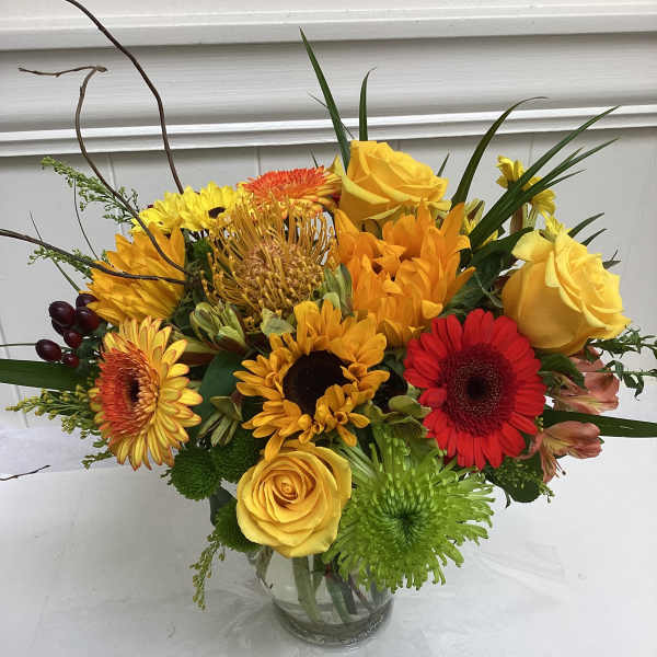 Bright mixed bouquet of yellow roses, sunflowers, red gerberas and green mums in a clear glass vase