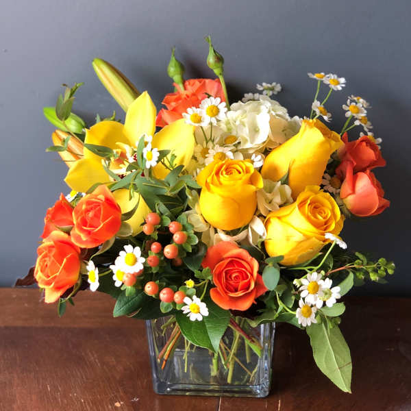 Bright arrangement of yellow roses, orange roses, lilies, and white daisies in a clear glass cube vase