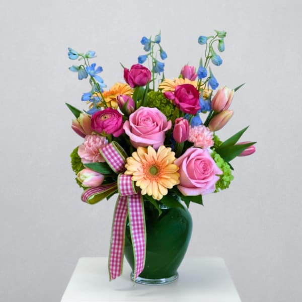 Colorful bouquet of pink roses, tulips, gerbera daisies, and blue delphinium in a green vase with ribbon.