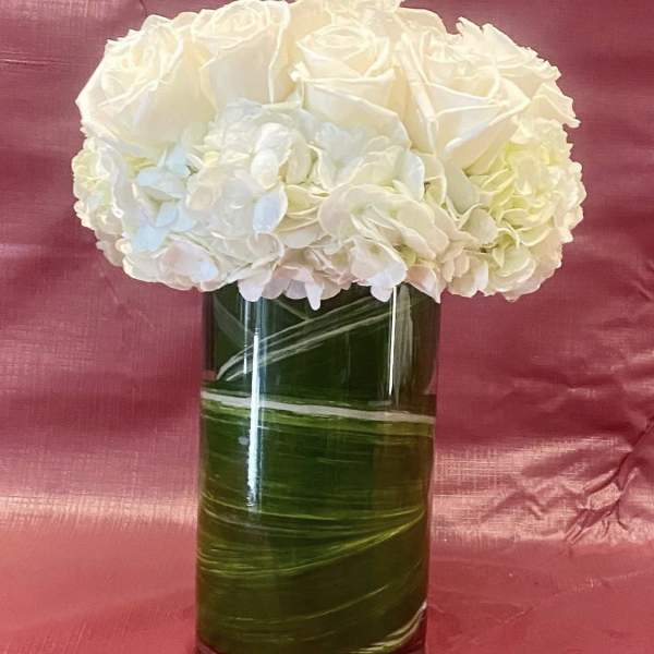 Custom arrangement of white roses & white hydrangeas in leaf lined clear glass vase.