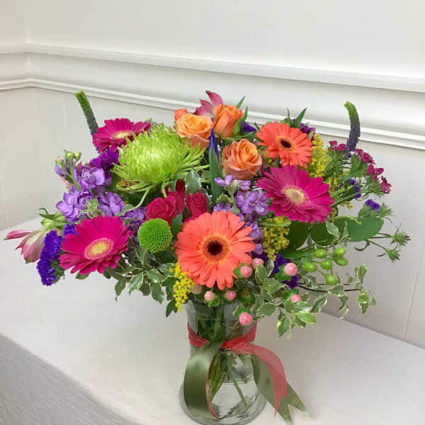 Vibrant mixed bouquet of orange roses and gerbera daisies with purple and green blooms in a clear glass vase