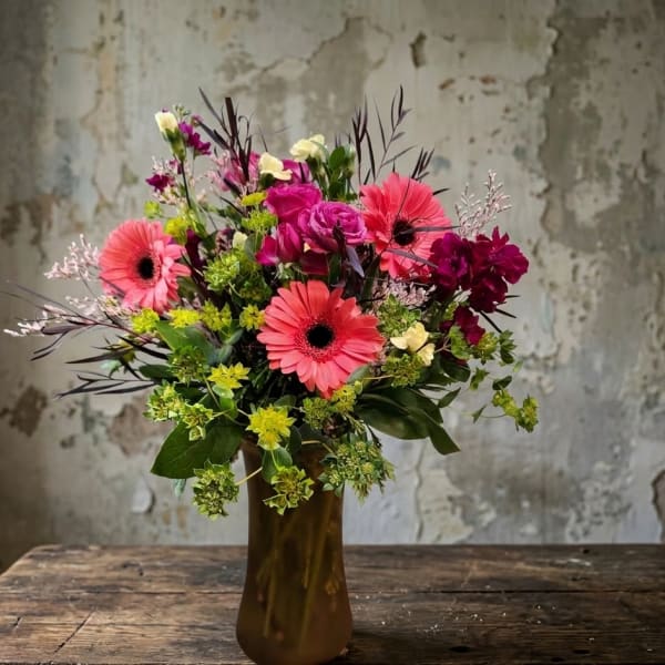 Tall bouquet of pink gerbera daisies and mixed blooms in an amber glass vase on a rustic wooden table.