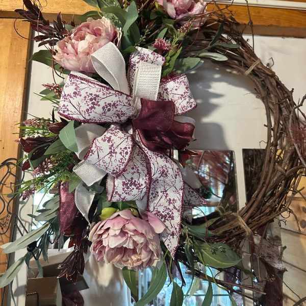 Grapevine wreath with pink peony blooms, greenery, and large white and burgundy patterned bow.