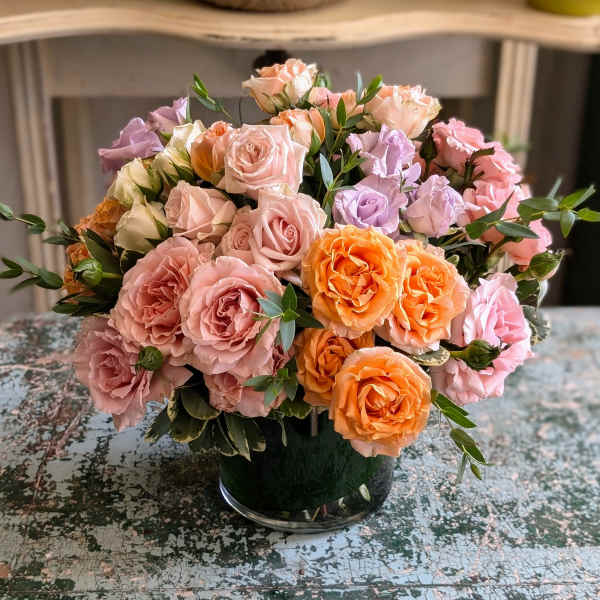 Low round arrangement of peach, pink, orange, and lavender roses in a clear glass vase