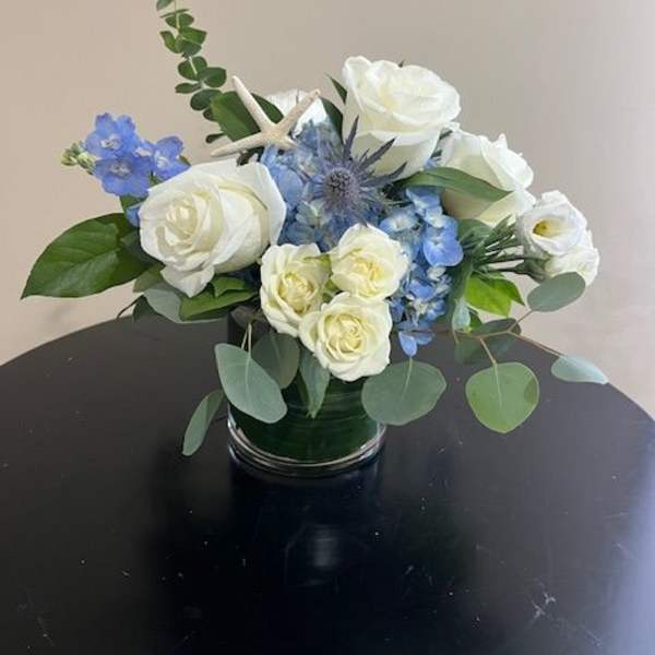Low arrangement of white roses and blue flowers in a clear glass vase with a small starfish accent.