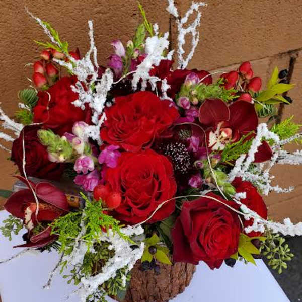 Low arrangement of red roses, orchids, and pink blooms with white frosted accents in a rustic bark container