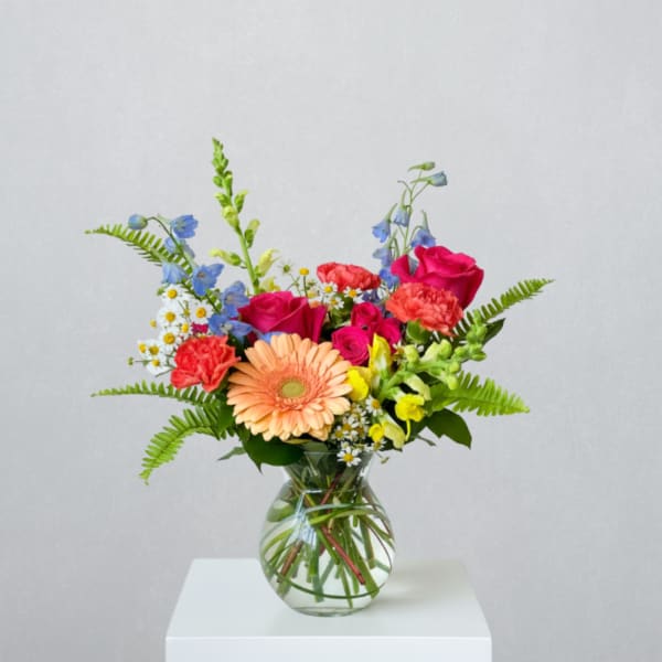 Mixed bouquet of bright pink, peach, yellow, and blue flowers in a clear glass vase.