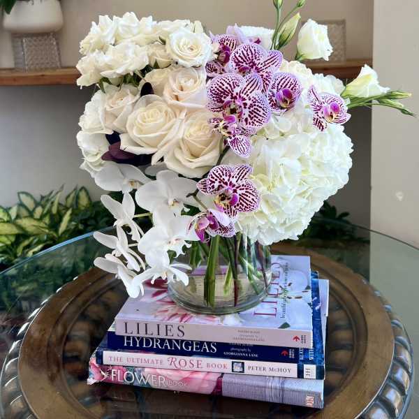 White roses, hydrangeas, and cascading orchids with purple speckled blooms in a clear glass vase on stacked books.