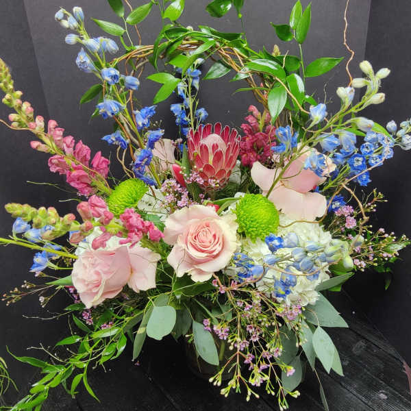 Pink roses and blue flowers arranged in a black container with a leafy arch