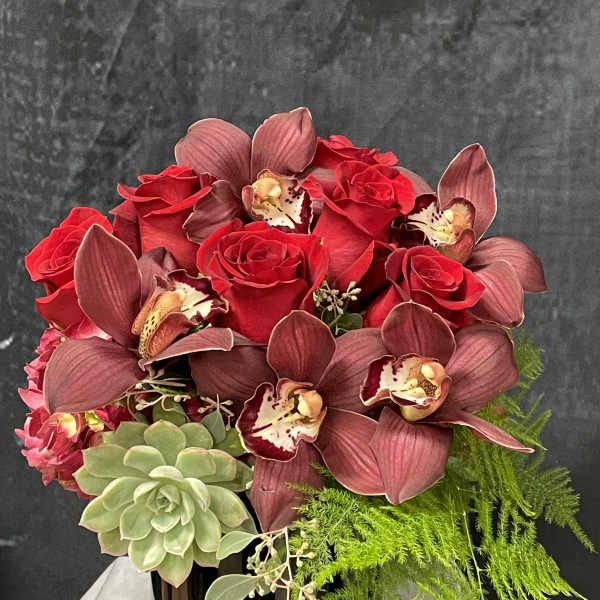Compact arrangement of red roses, burgundy orchids, and succulents in a black vase
