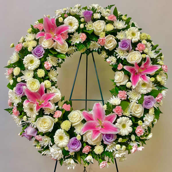 Circular floral wreath with pink lilies, white roses, and purple roses on a stand