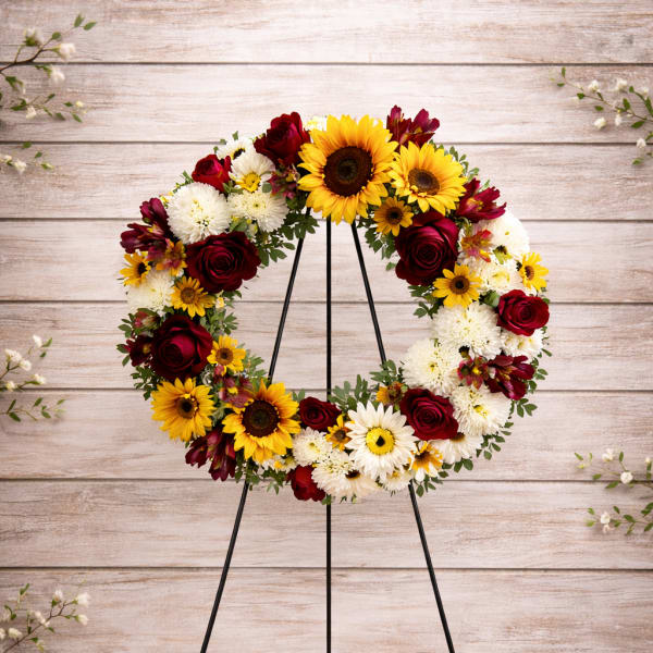 Circular wreath of sunflowers, red roses, and white daisies on a black stand