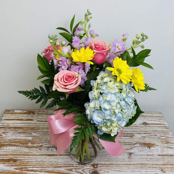 Mixed bouquet of pink roses, yellow daisies, blue hydrangea and lavender stems in a clear vase with pink ribbon