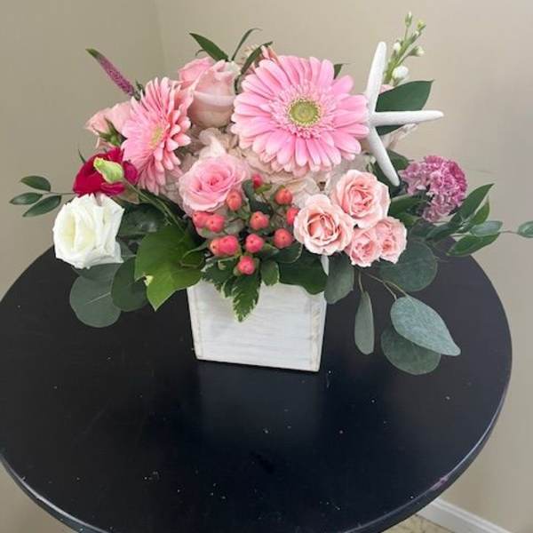 Pink gerbera daisies, roses, and spray roses in a white wooden cube with a decorative starfish