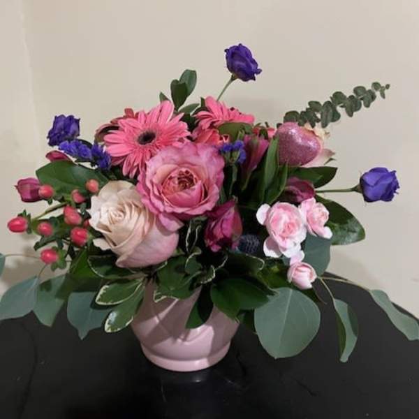 Pink and purple flower arrangement with roses and gerbera daisies in a pale pink ceramic bowl.