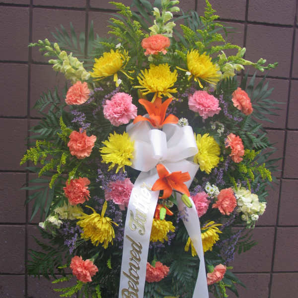 Standing funeral wreath with yellow and coral flowers and a white ribbon