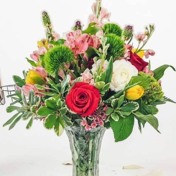 Mixed bouquet of red roses, yellow tulips, white blooms, and pink flowers in a clear glass vase