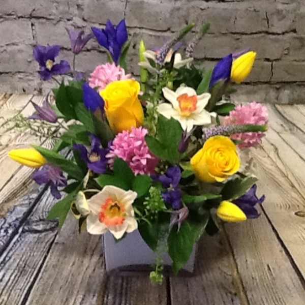Bright mixed arrangement with yellow roses, tulips, daffodils, pink hyacinths and purple flowers in a square vase