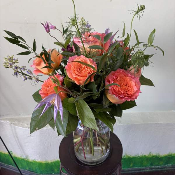 Vase arrangement of coral roses with purple accent flowers in a clear glass vase