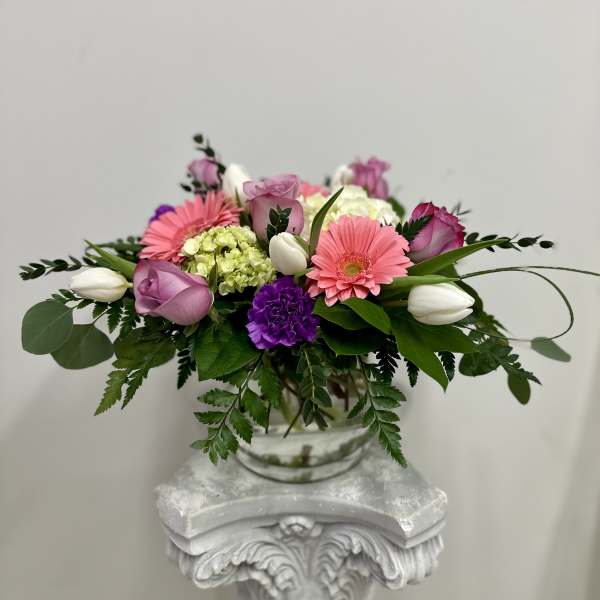 Low glass vase arrangement with pink gerbera daisies, purple roses, white tulips, and green hydrangea