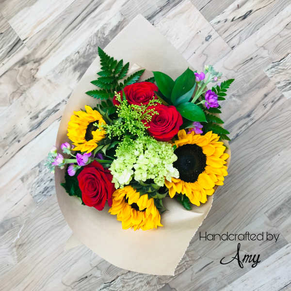 Wrapped bouquet of yellow sunflowers, red roses, hydrangea, and purple blooms on a wood background