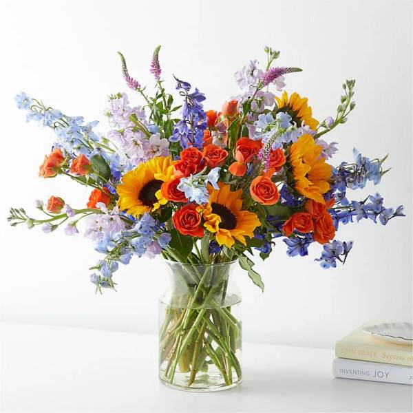 Bouquet of sunflowers, orange roses, and blue flowers in a clear glass vase