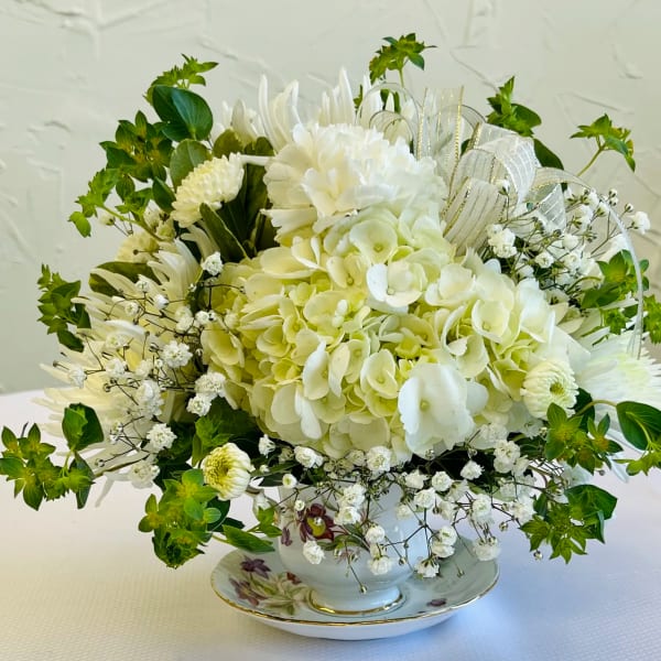 White floral arrangement in a teacup-style vase with green accents and ribbon