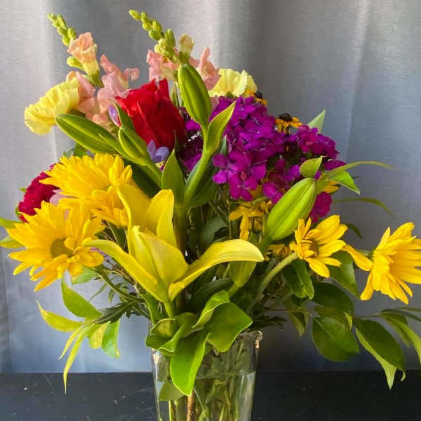 Mixed bouquet of yellow daisies, lilies, and purple flowers in a clear vase