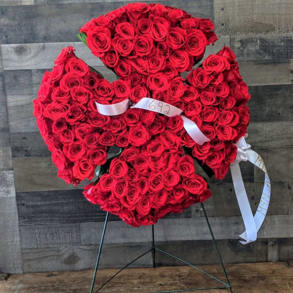 Standing spray of densely packed red roses in a cross-like shape on an easel with white ribbons.