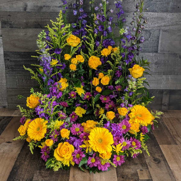 Tall one-sided arrangement of yellow roses and gerbera daisies with purple delphinium and daisy mums