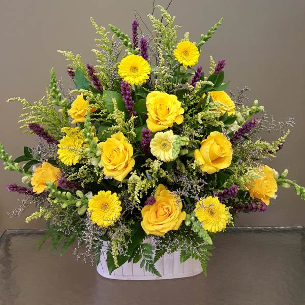 Large yellow rose and gerbera daisy arrangement with purple accents in a white basket.
