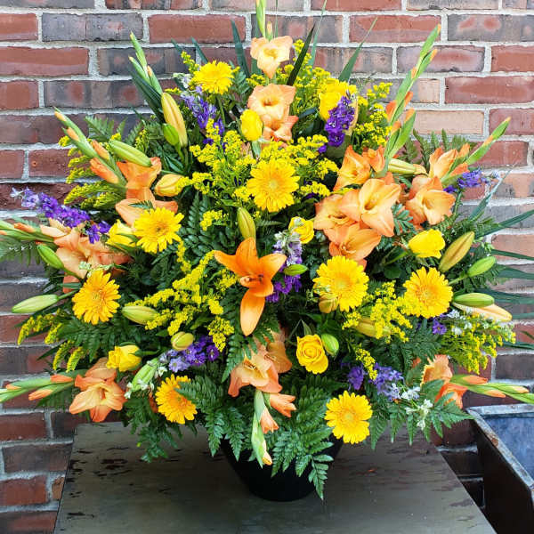 Tall arrangement of yellow gerbera daisies, orange lilies, and mixed flowers in a black urn.