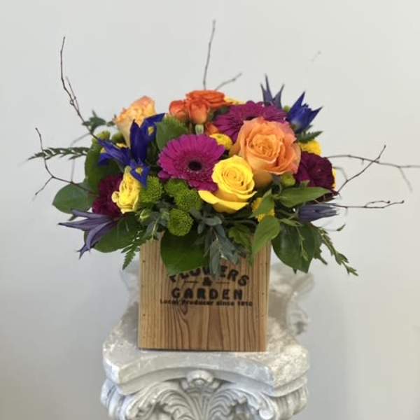 Bright mixed arrangement of yellow and orange roses, purple gerbera and blue iris in a wooden box.