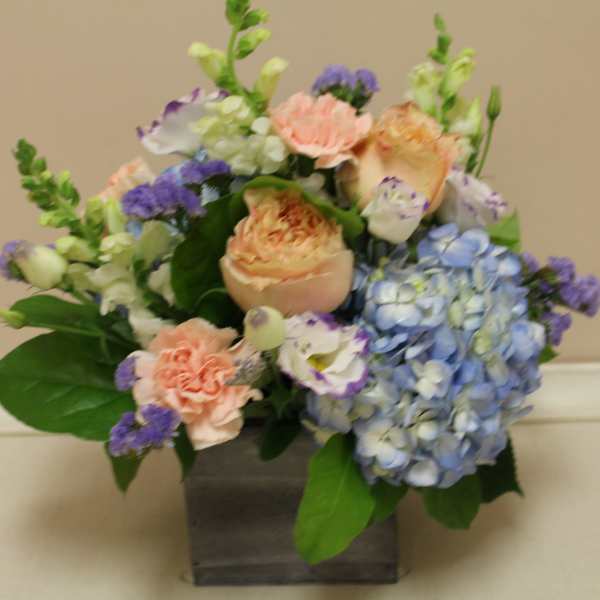 Mixed bouquet with blue hydrangea, peach roses, and purple accents in a square vase