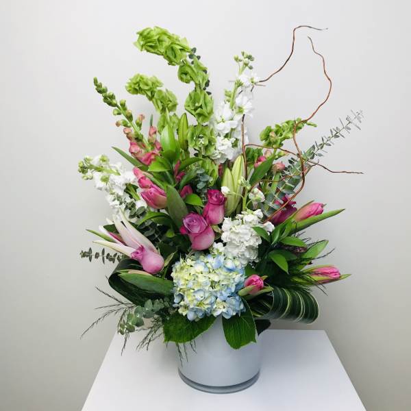Tall arrangement of pink roses and tulips with green Bells of Ireland, white lilies, and blue hydrangea in a white vase