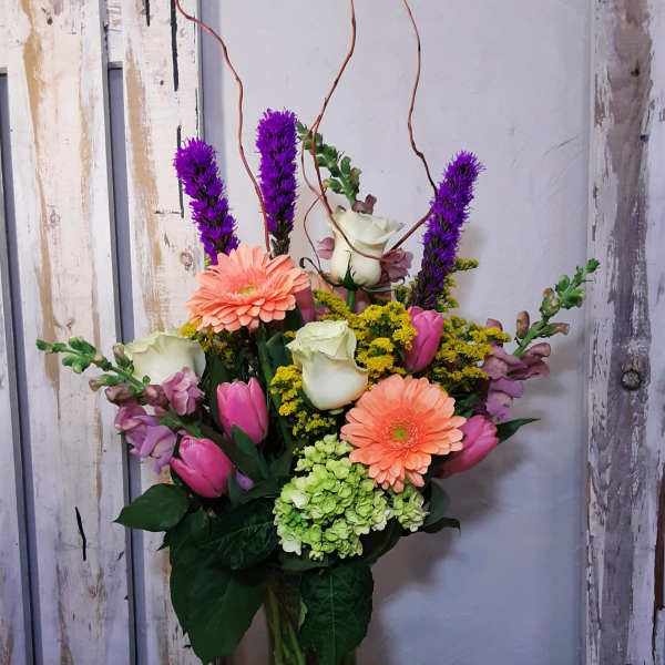 Tall mixed arrangement of peach gerberas, pink tulips, white roses, and purple flowers in a clear glass vase