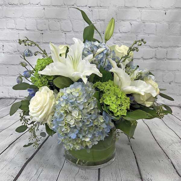 Low glass vase with white lilies, roses, blue hydrangeas, and bright green blooms in a rounded arrangement