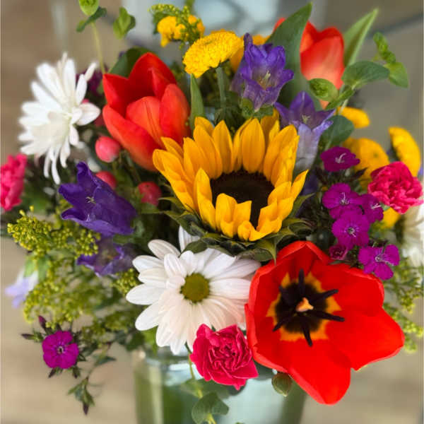 Colorful mixed bouquet with a sunflower, tulips, daisies, and purple blooms in a glass vase