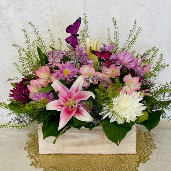 Pink lilies and purple daisies arranged in a white wooden box with butterfly picks.