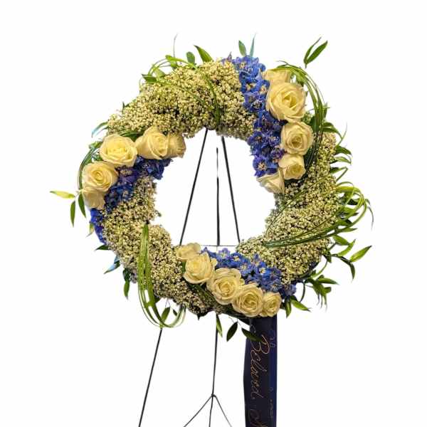 Standing wreath of cream roses, blue blooms, and white filler flowers on an easel with a dark ribbon