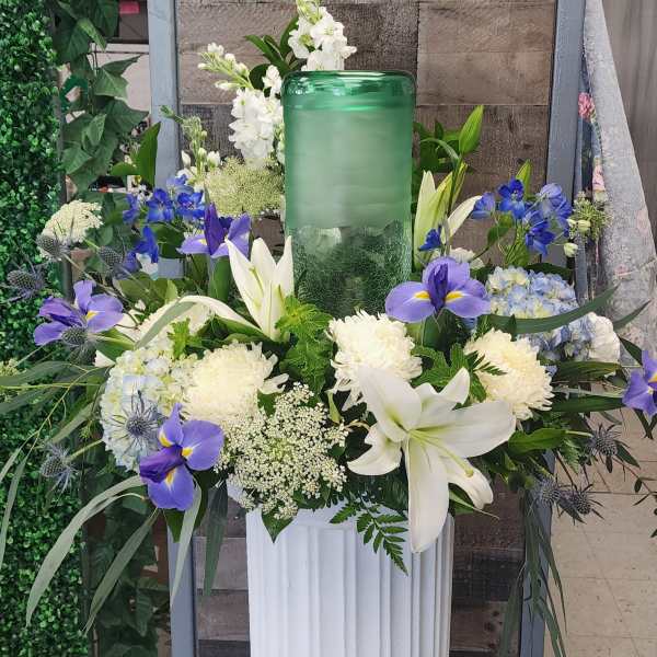 White lilies and blue-purple flowers arranged around a green glass candle holder